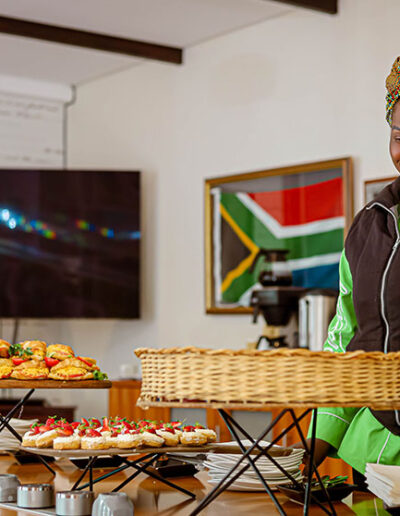 Woman in traditional attire arranging pastries on buffet table with South African flag in background