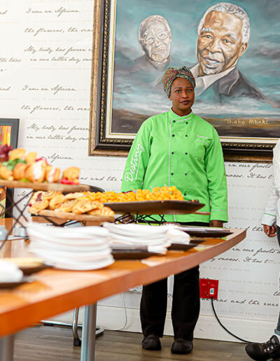 Chef in bright green coat standing near a table of finger foods and framed artwork, ready to deliver high-end event catering with a local cultural touch