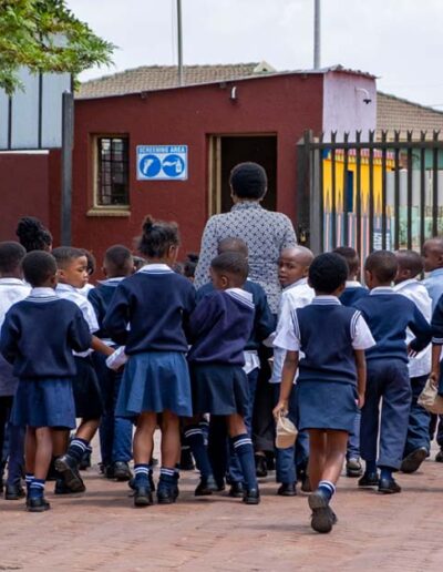 Children in school uniforms walking towards the school entrance under teacher supervision.