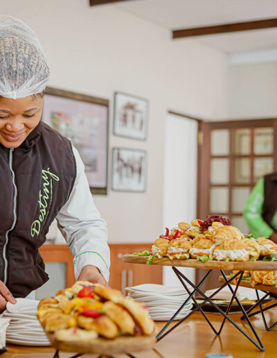 Catering staff in Destiny uniform arranging plates of sandwiches and pastries on a buffet table.